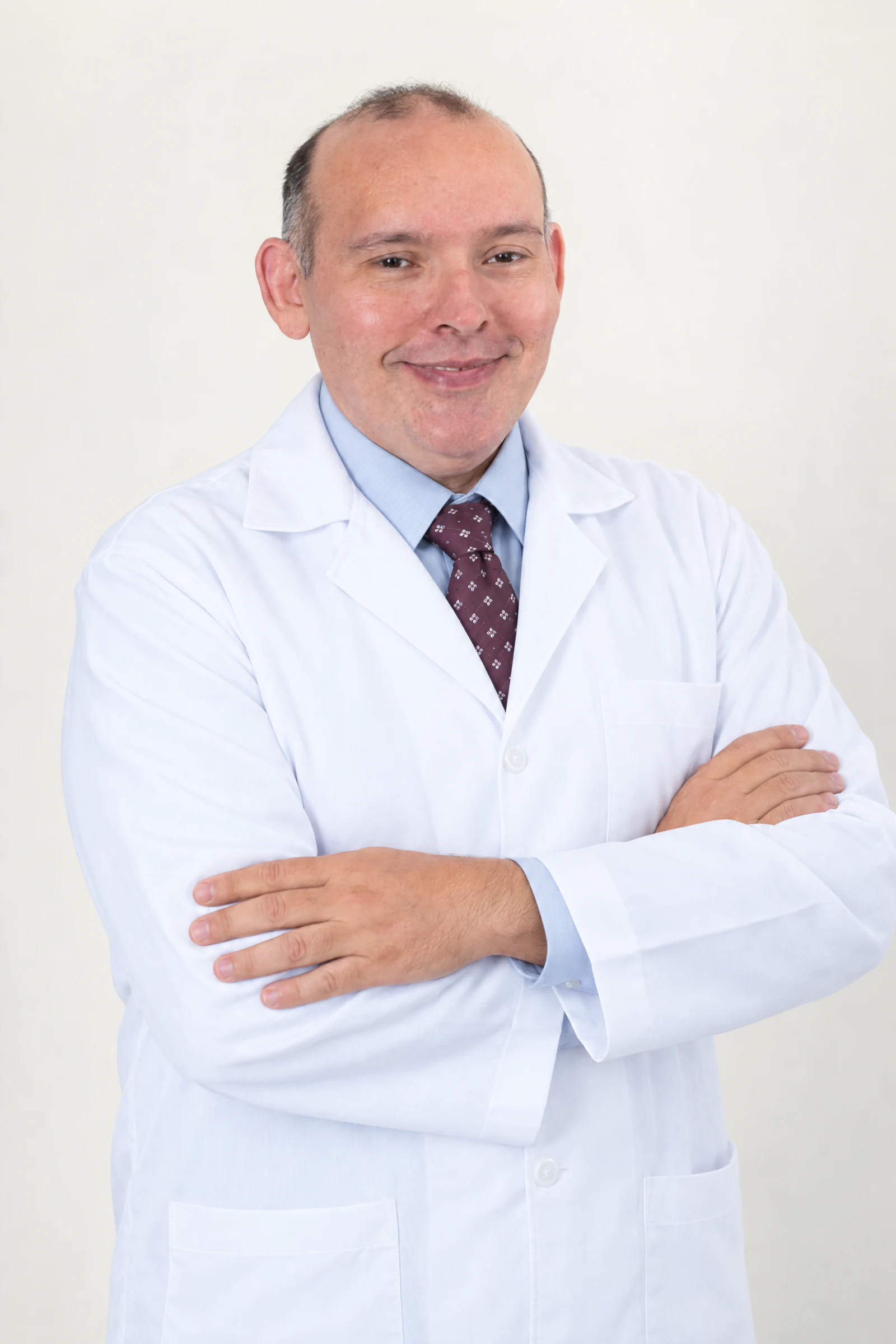 Dr. Avelino Júnior de jaleco branco, camisa azul-clara e gravata, com os braços cruzados